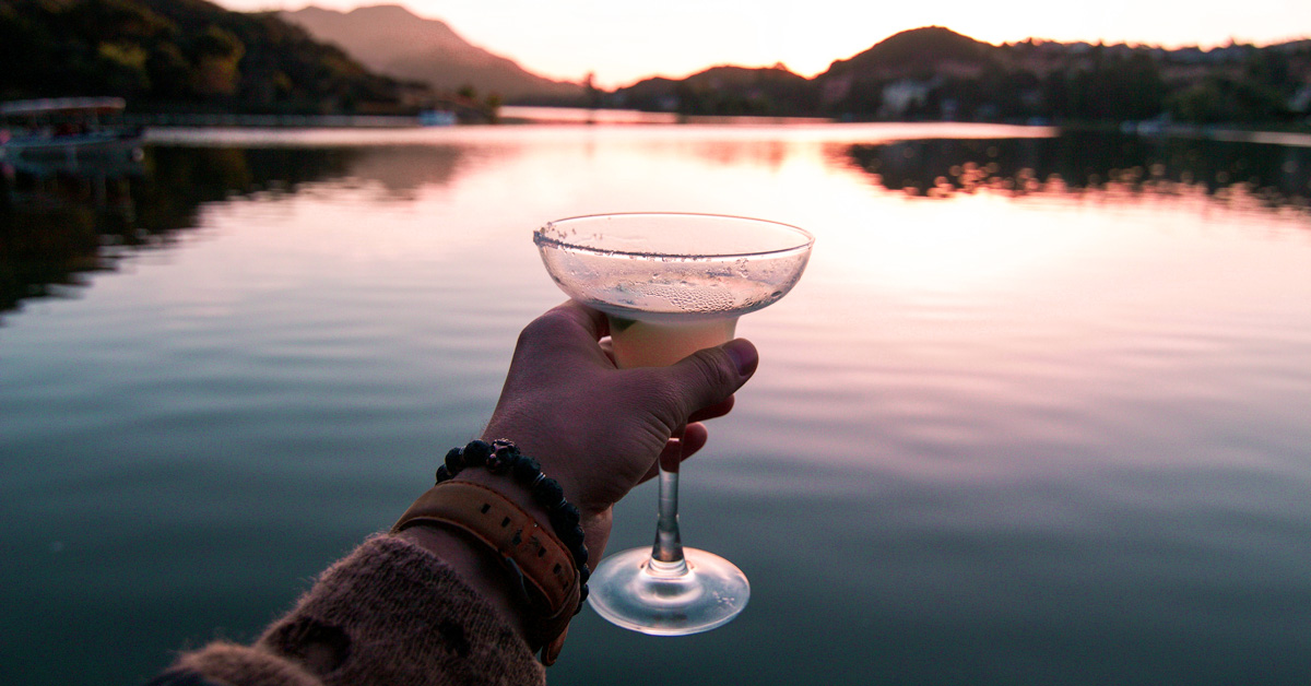 Une main tient un verre de punch devant un paysage de lac.